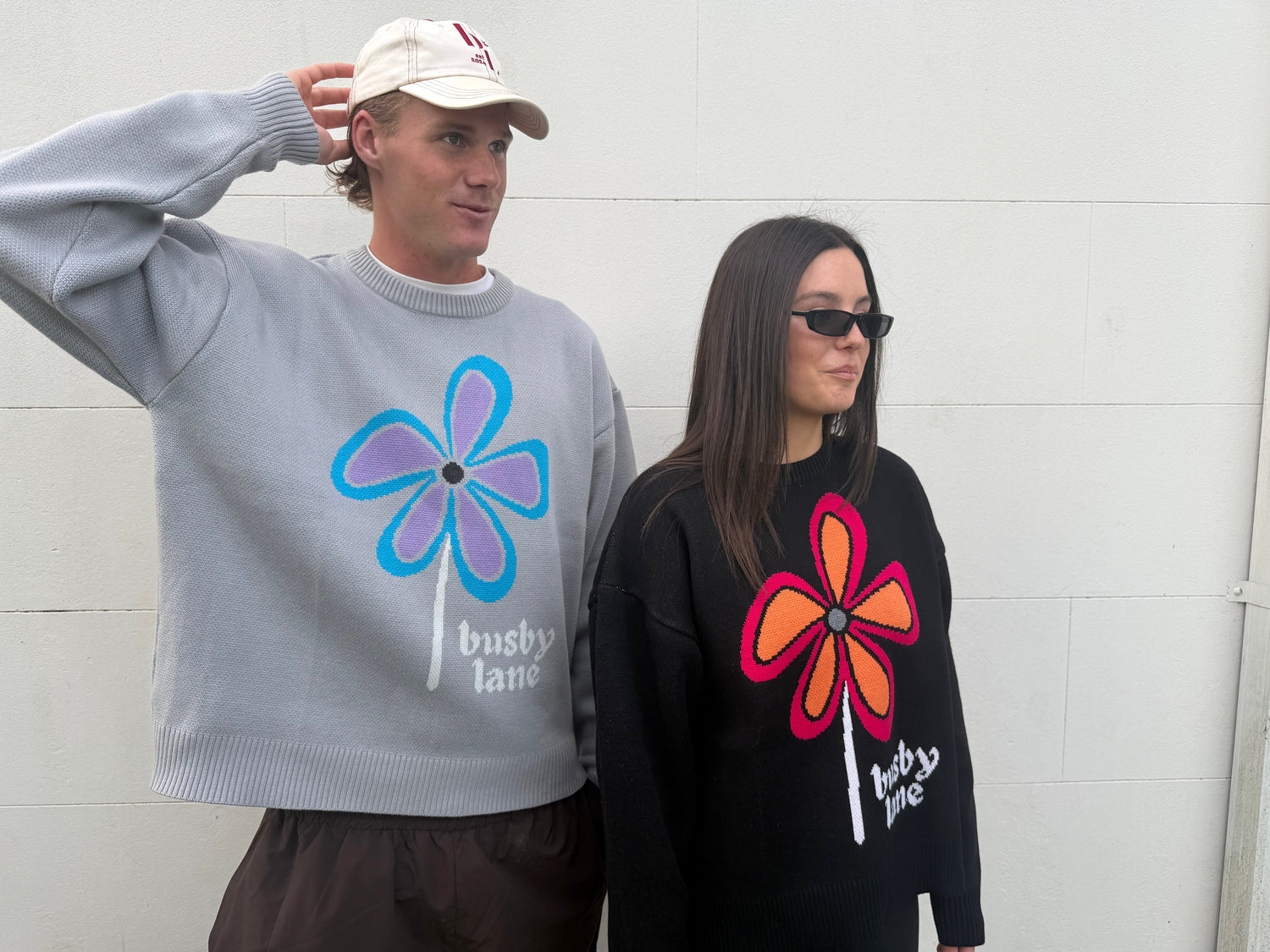 Two models wearing oversized crewneck sweaters with large flower graphics and 'busby lane' text, standing against a white wall.